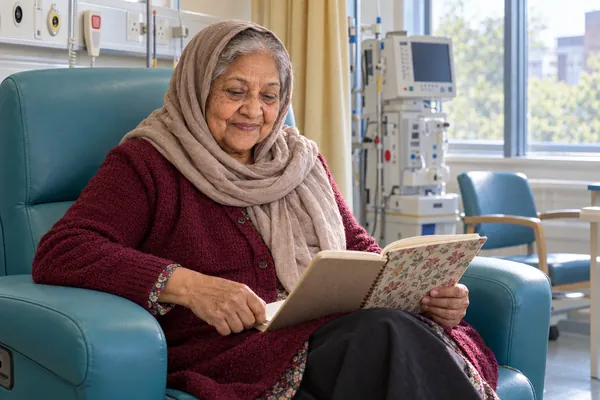 Wygenerowany przez AI wizerunek postaci Amina Begum, wiek: 74, w fikcyjnym brytyjskim szpitalu. Amina czyta zeszyt w turkusowym rozkładanym fotelu oddziału dziennego, w szarobrązowej chuście i bordowym kardiganie.